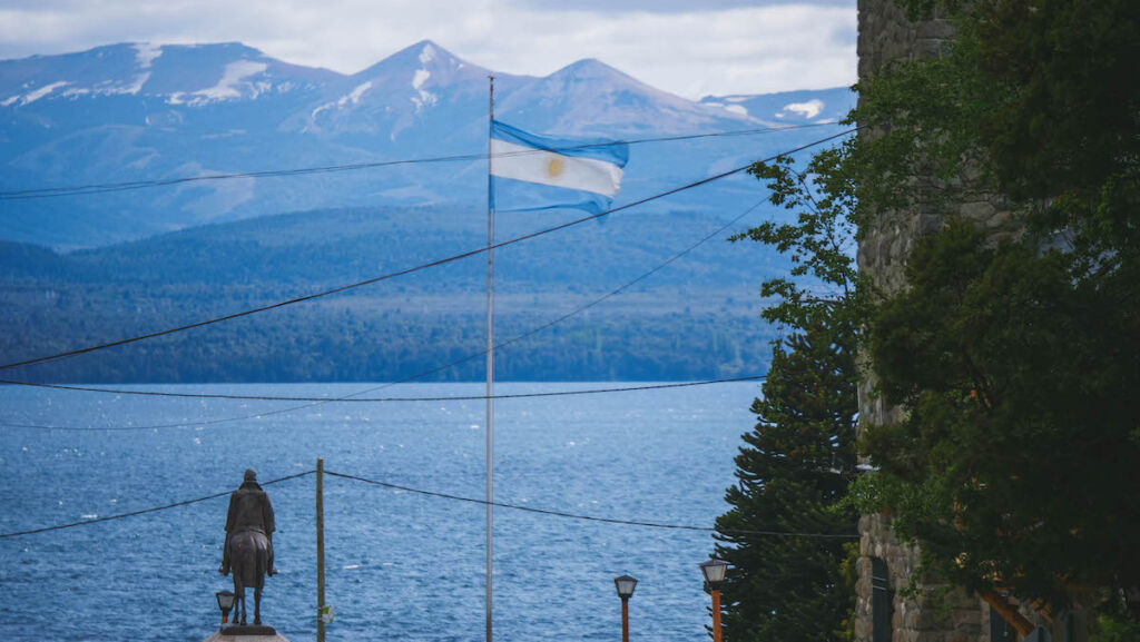 An Argentine flag blows in the wind in downtown Bariloche with lake and mountain views in the horizon. This is a typical scene you can see on a Bariloche walking tour. 