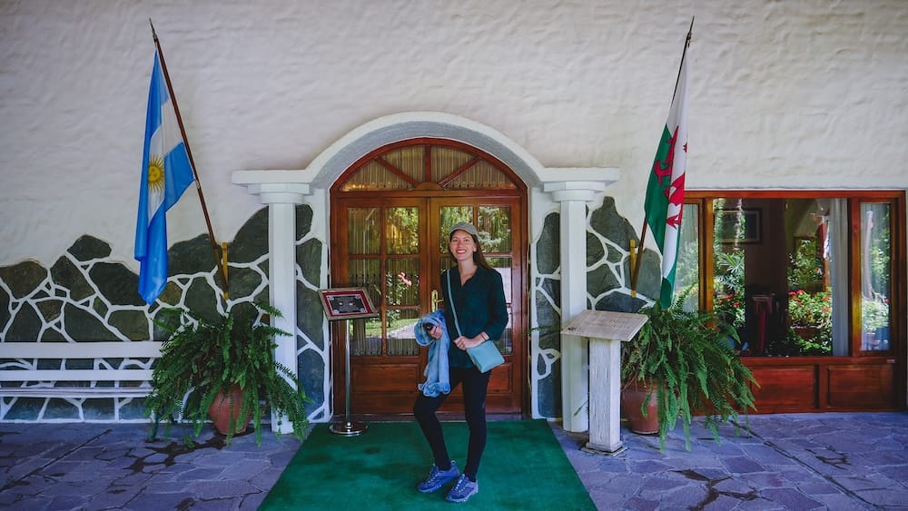 Audrey standing in front of the Ty Te Caerdydd tea house in Gaiman with an Argentine flag and a Welsh flag.