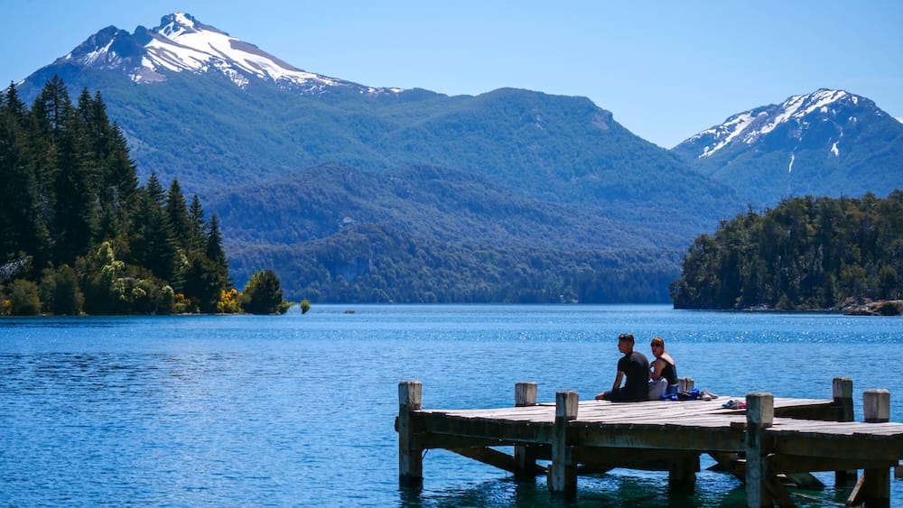 Two tourists sitting on the wooden dock at Playa de los Toros on a day trip to Isla Victoria, Bariloche.